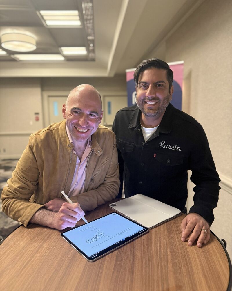 Grindr CEO George Arison and Freddie CEO Dr. Husein Moloo at a table with a signed agreement on a tablet